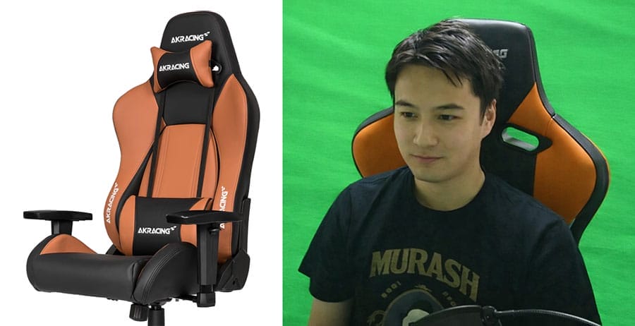 Japanese streamer Kato Junichi sitting in an orange-and-black AKRacing Premium gaming chair during a livestream, with a green screen behind him.