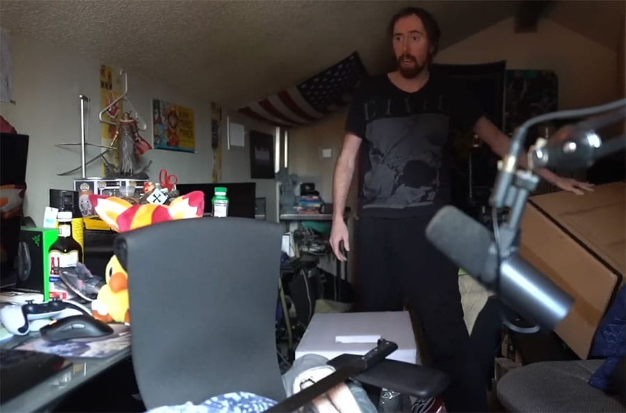 ZackRawrr standing in a cluttered streaming room with a Herman Miller Embody chair visible at his desk, surrounded by gaming equipment, boxes, and a microphone setup.